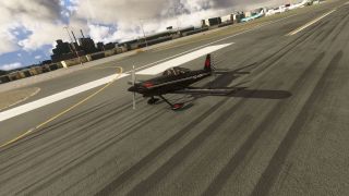 A black single-engine aircraft with a checkered tail sits on a tire-marked runway, with terminal buildings and parked airliners behind it.