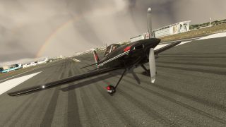 A black single-engine propeller aircraft sits on a marked runway, with a rainbow, parked airliners, and airport buildings behind it.