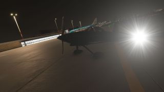 A propeller aircraft sits on a floodlit apron at night, with bright light glare and a KLM jet behind it.