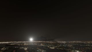A single-engine airplane flies above illuminated city streets and waterways at night, with a bright light shining from its left wing.
