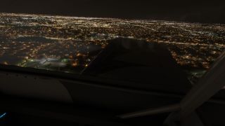 An aircraft wing fills the side window view above a sprawling city illuminated by streetlights and building lights at night.
