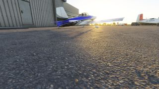 A blue-and-white light aircraft sits beside a corrugated hangar, with low sunlight casting long shadows across the apron.