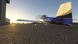 A blue, black, and white taildragger marked EC-KCN sits beside hangars with its propeller spinning in low sunlight.