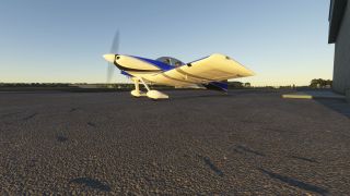 A blue-and-white low-wing airplane sits on the apron beside a hangar, with its propeller spinning and two occupants visible.
