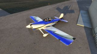 A blue-and-white low-wing aircraft with two occupants sits beside a hangar, casting a long shadow across the apron.