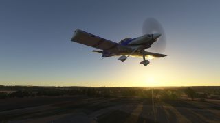 A blue and white propeller aircraft with fixed landing gear flies low over fields and trees with the sun near the horizon.
