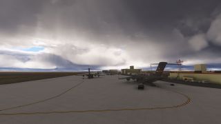 Military transport aircraft sit on a concrete apron beside airport buildings and service vehicles beneath dense, dark clouds.