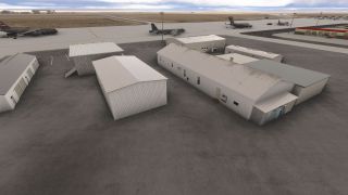 Several military transport aircraft sit on a broad apron behind corrugated metal airport buildings, with taxiways and distant mountains beyond.