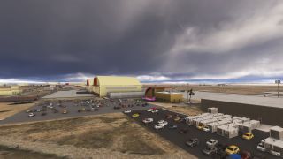 Large yellow aircraft hangars stand beside crowded parking lots and rows of portable buildings beneath dark, low clouds.