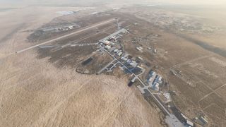 An aerial view shows a desert airfield with a long runway, connecting taxiways, scattered hangars, and aircraft parked on aprons.