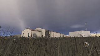 Low utility buildings with metal doors stand beyond tall grass and wildflowers beneath dark clouds, with a floodlight mast nearby.