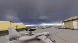 A U.S. Air Force radar aircraft sits on an apron between yellow hangars beneath dark clouds, with other military aircraft nearby.