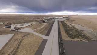 A broad taxiway leads toward hangars and parked aircraft, with cylindrical storage tanks and barren terrain beneath dark clouds.