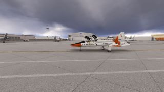 A white and orange U.S. Air Force jet sits with both canopies open on an apron beneath dark clouds, near NASA hangars.