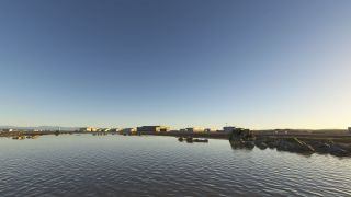 Airport hangars line a rocky shoreline beside rippled water, with distant hills beneath a clear blue sky.