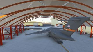A gray military transport aircraft sits beneath red arched hangar beams, with its rear cargo ramp lowered and Edwards tail markings visible.