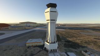 A tall, angular control tower stands beside airport taxiways and hangars, with parked aircraft and dry terrain in the background.
