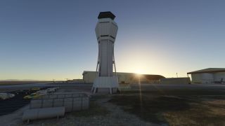 A tall airport control tower stands beside parked vehicles, with hangars and aircraft behind it under a low sun.