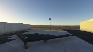 A dark flying-wing bomber sits with landing gear extended on a concrete apron between hangars, with a floodlight tower behind it.