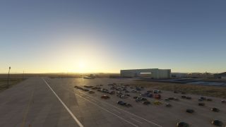 Rows of cars occupy a broad airport apron beside hangars, with a low sun casting long shadows across the pavement.