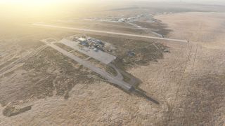 Two paved runways, connecting taxiways, hangars, and parked aircraft occupy an airfield surrounded by dry terrain under low sunlight.