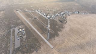 An airport in arid terrain has intersecting runways, connecting taxiways, scattered hangars, and several small aircraft parked on an apron.