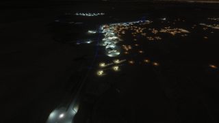 An airport seen from above at night has illuminated hangars and aprons, blue taxiway lights, and scattered amber ground lighting.