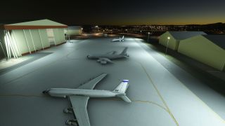Three U.S. Air Force jets sit on a floodlit apron between hangars, with yellow taxiway markings and distant lights beneath a dark sky.