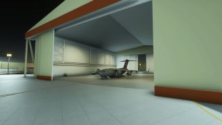 A gray four-engine military transport aircraft sits inside an open, illuminated hangar at night, with boarding stairs beside its nose.