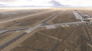 An aerial view shows a desert airport with parallel runways, connecting taxiways, hangars, and parked aircraft on a large apron.