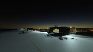 Large transport aircraft sit on a floodlit apron beside hangars, with ground vehicles nearby and blue taxiway lights stretching into the darkness.