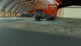 A gray military transport aircraft sits inside an orange-framed arched hangar, with a green heavy truck parked in front.