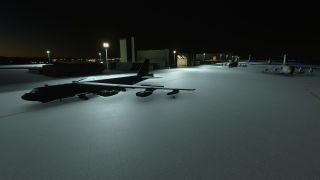 A B-52 bomber sits on a floodlit apron at night, with hangars and military transport aircraft behind it.