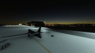A four-engine bomber sits on a floodlit apron at dusk, with hangars ahead and ground service equipment nearby.