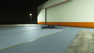 A dark, long-winged aircraft sits on a floodlit apron beside a large white hangar with orange trim at night.