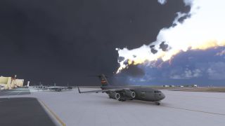A gray U.S. Air Force C-17 sits on an apron beneath dark clouds, with parked aircraft and hangars behind it.