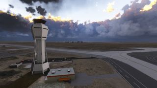 An angular airport control tower stands beside intersecting taxiways, parked cars, and a low building beneath dark, sunlit clouds.