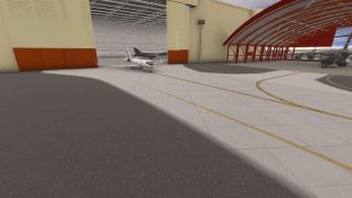 A white business jet sits outside an open hangar beside gray military transport aircraft sheltered beneath a red steel canopy.