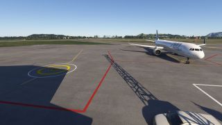 An Airbus jet sits on the apron beside red boundary lines and a circular helipad marking, with wooded hills beyond.