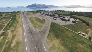 A coastal airport has a single runway, connecting taxiways, and parked aircraft beside terminal buildings, with water and mountains beyond.