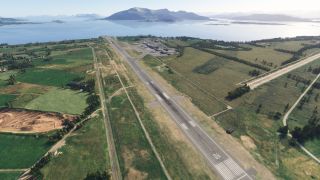 Runway 06 extends through green fields toward waterfront airport buildings, with parked aircraft, distant mountains, and a bridge across the water.