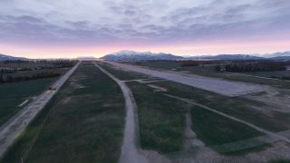 A runway and connecting taxiways cross grassy terrain, with snow-covered mountains beneath a pink horizon and layered clouds.