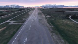 A runway with displaced threshold arrows extends past grassy fields toward water and snow-covered mountains beneath a pink sky.