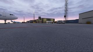 An airliner’s nose overlooks an airport apron with service vehicles, cargo buildings, and lattice lighting towers beneath a cloudy pink sky.