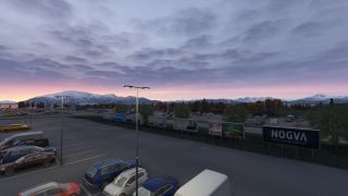 Parked cars, illuminated lampposts, and roadside billboards fill the foreground beneath snow-covered mountains and a cloudy pink sky.