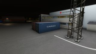 Blue and red cargo containers sit outside a Lufttransport building at night, beside a metal lattice tower and white pavement markings.