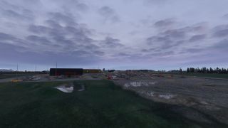 A large parking lot contains rows of cars beside low buildings, with grassy drainage channels in the foreground beneath overcast skies.
