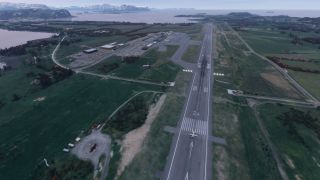 A small aircraft sits near runway 24’s threshold at a coastal airport, with terminal buildings, green fields, and snow-covered mountains beyond.