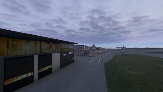 Airport service buildings and vehicles line a marked apron road, with parked airliners and a control tower beneath overcast skies.