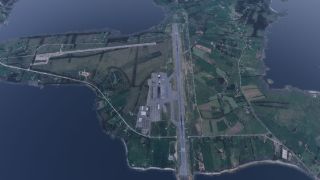 An airport occupies a narrow peninsula, with a long runway, connecting taxiways, parked aircraft, and surrounding fields bordered by water.